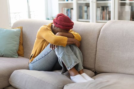 Photo for Depressed unhappy frustrated young african american woman wearing red turban sitting on couch with legs up at home, suffering from loneliness, hugging her knees and crying, copy space - Royalty Free Image