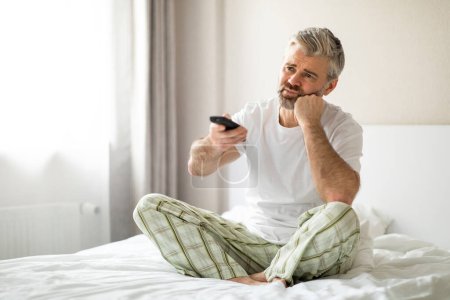 Photo for Bored unhappy middle aged man sitting on bed, switching channels on tv, touching his face, looking for something interesting, wearing pajamas, spend weekend at home alone, copy space - Royalty Free Image