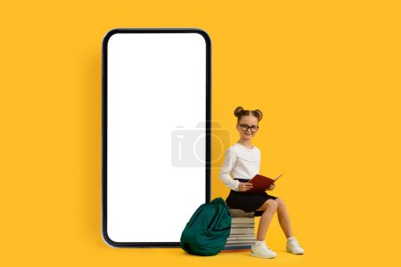 Photo for Study App. Cute little schoolgirl sitting on pile of books near huge blank smartphone with white screen over yellow studio background, happy female pupil recommending educational application, mockup - Royalty Free Image