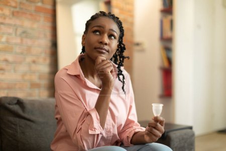 Photo for Pensive black woman holding reusable menstrual cup, effective safe affordable menstrual product, good protection from leakage, thinking how to use it, home interior - Royalty Free Image