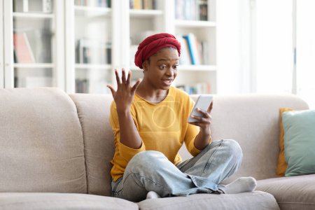 Foto de Sorprendido joven mujer negra feliz en traje casual y turbante rojo sentado en el sofá, usando el teléfono celular en casa, usuario femenino mirando la pantalla del gadget y el gesto, ganó la lotería, espacio de copia - Imagen libre de derechos