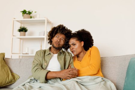 Téléchargez les photos : Couple afro-américain anxieux regardant un film effrayant ou des nouvelles négatives à la télévision embrassant assis sur le canapé ensemble dans le salon moderne à la maison. Concept de crise et de stress - en image libre de droit