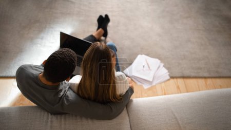Photo for Millennial european woman hugs arab guy, watch video on laptop with empty screen, works with documents, rests in living room, panorama, top view. Home accounting, pay taxes, new normal and gadget - Royalty Free Image