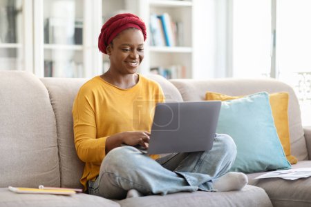 Téléchargez les photos : Jolie femme afro-américaine millénaire joyeuse avec turban rouge en tenue décontractée assise sur le canapé, tapant sur le clavier de l'ordinateur portable pc et souriant, nomade numérique féminine travaillant de la maison, espace de copie - en image libre de droit