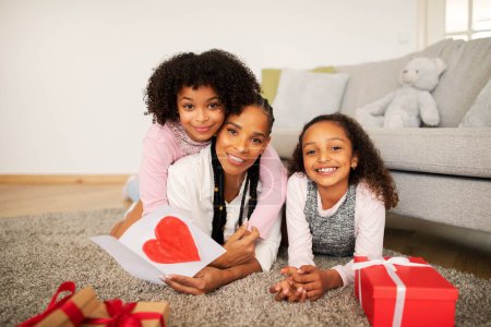 Téléchargez les photos : Aimer maman noire et ses filles célébrer les vacances de la fête des mères ensemble à la maison. Maman tenant la carte avec le coeur posant avec deux filles d'enfant allongé parmi enveloppé présent boîtes souriant à la caméra - en image libre de droit