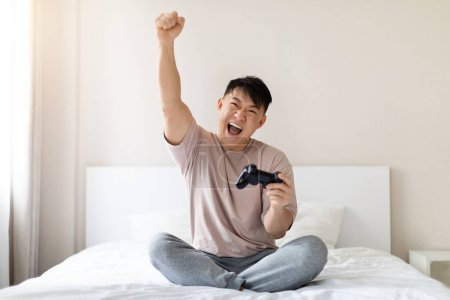 Photo for Resting at home, domestic entertainment concept. Happy emotional middle aged asian man sitting on bed at home, holding joystick and raising hand up, playing video games, copy space - Royalty Free Image