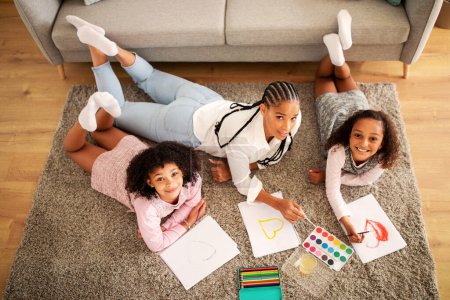 Foto de Feliz madre negra y dos hijas dibujando juntas acostadas en el piso en la sala de estar moderna en casa el fin de semana, sonriendo a la cámara. Hobby familiar y ocio. Vista superior Shot - Imagen libre de derechos