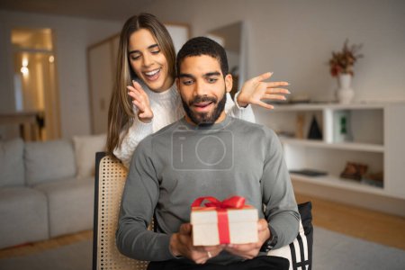Photo for Smiling millennial european female opens eyes with hands to arab husband, gives box with present in living room interior. Surprise for birthday, anniversary and holiday, love, relationships at home - Royalty Free Image