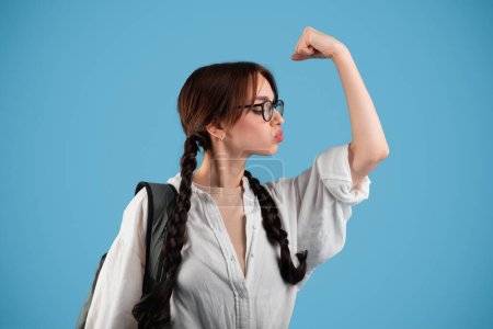 Photo for Happy european teenager girl student with pigtails and backpack in glasses kisses biceps muscle isolated on blue studio background. Female strength power, great test result, fun, study and education - Royalty Free Image
