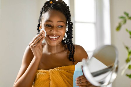 Téléchargez les photos : Soin du visage. Heureuse belle femme afro-américaine en serviette regardant miroir et en utilisant de l'eau micellaire avec coton pour le démaquillage, intérieur de la maison, espace libre - en image libre de droit