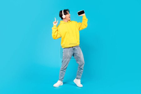 Photo for Korean Teen Boy Singing Holding Smartphone Like Microphone Wearing Earphones Having Fun Standing On Blue Studio Background. Great Musical Application Advertisement. Full Length Shot - Royalty Free Image