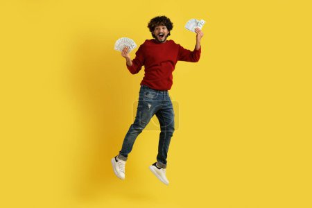 Photo for Happy lucky rich millennial hindu guy in stylish casual outfit jumping in the air with cash dollar banknotes in his hands, celebrating success, yellow studio background. Lottery, bet, gambling - Royalty Free Image