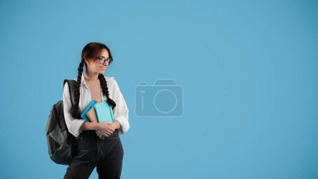 Photo for Cheerful european smart teenager girl with pigtails and backpack in glasses hold books, looks at empty space, isolated on blue studio background. Modern study, knowledge and education, ad and offer - Royalty Free Image