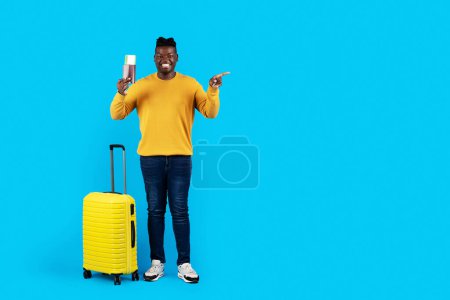Téléchargez les photos : Heureux homme afro-américain avec valise et passeport pointant vers l'espace de copie, voyageur afro-américain posant avec des bagages sur fond bleu studio, pleine longueur, espace de copie - en image libre de droit