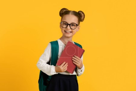 Photo for Education Concept. Portrait Of Smiling Cute Schoolgirl With Backpack And Books In Hands, Cheerful Little Female Pupil Wearing Eyeglasses Standing Isolated Over Yellow Studio Background, Copy Space - Royalty Free Image