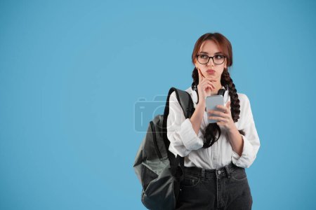 Photo for Pensive european teen girl student with pigtails, backpack in glasses thinks with phone, look at free space on blue studio background. Creating solution, got idea, app for study and education - Royalty Free Image