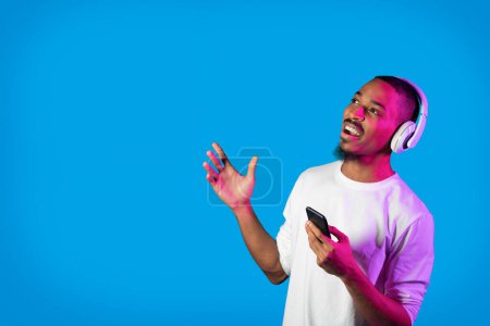 Téléchargez les photos : Joyeux jeune homme afro-américain excité en blanc manches longues écoutant de la musique en ligne, utilisant un casque sans fil, tenant un smartphone, regardant l'espace de copie sur fond de studio bleu sous la lumière néon - en image libre de droit