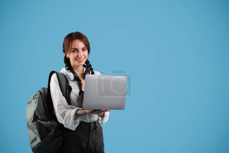 Téléchargez les photos : Étudiante adolescente européenne joyeuse avec des nattes et un sac à dos tapant sur ordinateur, bavardant sur les réseaux sociaux sur fond de studio bleu. App pour l'étude, la connaissance et l'éducation à distance - en image libre de droit