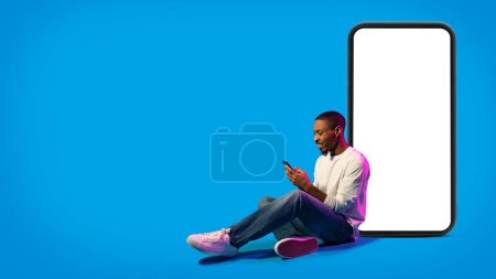Téléchargez les photos : Cool joyeux beau jeune homme noir en tenue décontractée assis sur le sol par un énorme smartphone avec écran blanc blanc sur fond bleu au néon, en utilisant un téléphone portable, une maquette, un espace de copie, une bannière - en image libre de droit