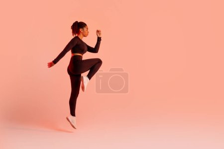 Foto de Ajuste atleta mujer saltando mientras hace ejercicio sobre fondo de estudio de neón melocotón, mirando a un lado en el espacio libre, longitud completa, vista lateral. Entrenamiento físico y concepto de motivación deportiva - Imagen libre de derechos