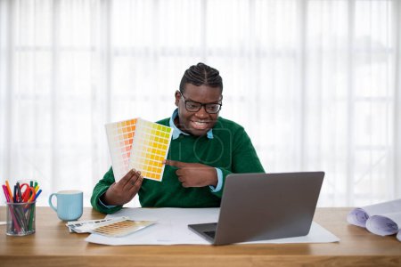 Téléchargez les photos : Heureux afro-américain mâle designer montrant palette de couleurs pour ordinateur portable ayant appel vidéo avec client assis au bureau moderne. Design et Couleurs Tendances Concept - en image libre de droit