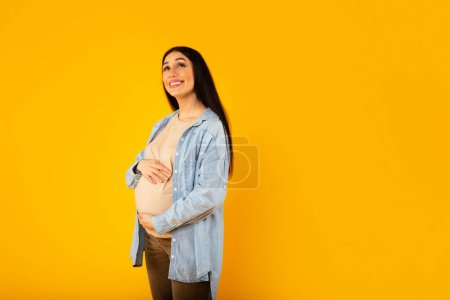 Téléchargez les photos : Femme enceinte souriante et rêveuse qui caresse le ventre enceinte en levant les yeux et en souriant, posant sur un fond de studio jaune avec un espace de copie gratuit - en image libre de droit
