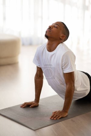Foto de Tiro franco vertical del joven atlético negro en ropa deportiva haciendo yoga en casa, chico afroamericano estirando el cuerpo en la alfombra de fitness en la mañana, espacio de copia, estilo de vida saludable y concepto de deporte - Imagen libre de derechos