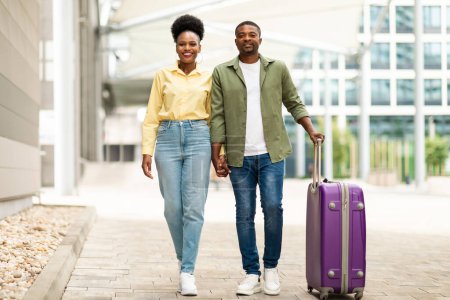 Téléchargez les photos : Couple de voyageurs noirs tenant la main Marcher avec une valise Aller en vacances près de l'aéroport moderne à l'extérieur. Conjoints voyageant ensemble Souriant à la caméra. Offre vols pas chers. Longueur totale - en image libre de droit