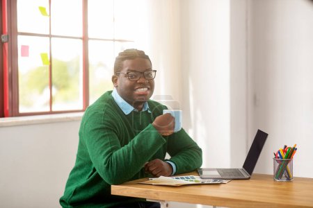 Foto de Diseñador masculino negro exitoso usando el ordenador portátil y disfrutando del café en el lugar de trabajo sentado en la oficina moderna, sonriendo a la cámara. Diseño Carrera y estilo de vida independiente Concepto - Imagen libre de derechos