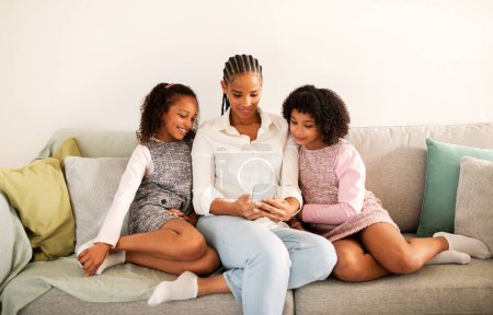 Photo for Cheerful Black Mother And Daughters Using Smartphone Together Browsing Internet And Texting Sitting On Couch At Home On Weekend. Family And Gadgets Lifestyle Concept - Royalty Free Image