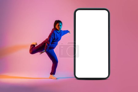 Foto de Bailarina profesional saltando cerca de un enorme smartphone con pantalla blanca en blanco, recomendando aplicación móvil con tutoriales de baile en línea, fondo de neón rosa, maqueta - Imagen libre de derechos