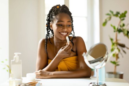 Foto de Feliz hermosa mujer negra usando bálsamo labial hidratante, señora sentada en la mesa de tocador, mirando al espejo pequeño, aplicando maquillaje desnudo luz todos los días en casa, espacio libre - Imagen libre de derechos