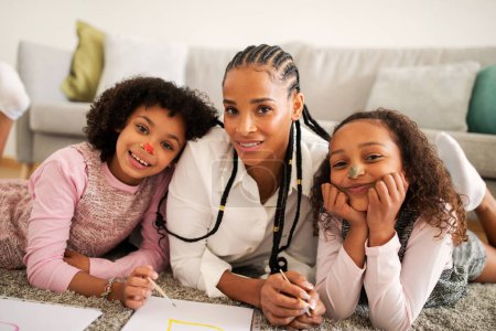 Téléchargez les photos : Loisirs en famille. Mère afro-américaine joyeuse et deux filles dessinant passer du temps ensemble allongé sur le sol souriant à la caméra à la maison. Bon week-end avec maman. Concept parental - en image libre de droit