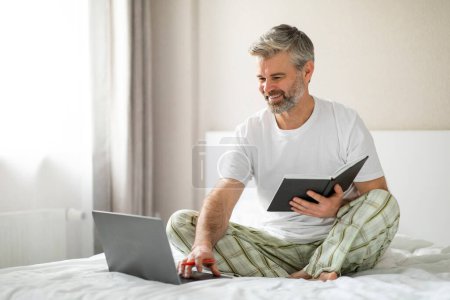 Téléchargez les photos : Enthousiaste positif beau aux cheveux gris homme mature portant un pyjama assis sur le lit à la maison, en utilisant un ordinateur portable PC moderne, prendre des notes dans un bloc-notes, assister à une formation en ligne ou webinaire, semaine de planification - en image libre de droit