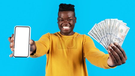 Photo for Big Profit. Cheerful Black Man Holding Dollar Cash Money And Blank Smartphone, Happy Young African American Guy Recommending Online Credit While Standing Isolated Over Blue Background, Mockup - Royalty Free Image