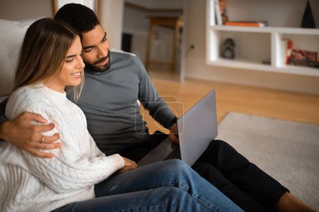 Photo for Smiling millennial arabic male hugs european female watch video on laptop, have meeting, enjoy spare time, sitting on floor in living room, close up. New normal and gadget, communication remotely - Royalty Free Image