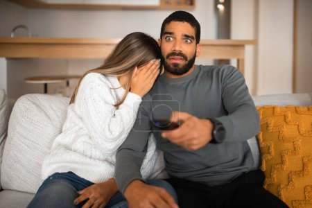 Photo for Shocked frightened millennial arab guy with beard, remote control and european woman close face, watch TV together on sofa in living room interior. Afraid, scared film at home and people emotions - Royalty Free Image