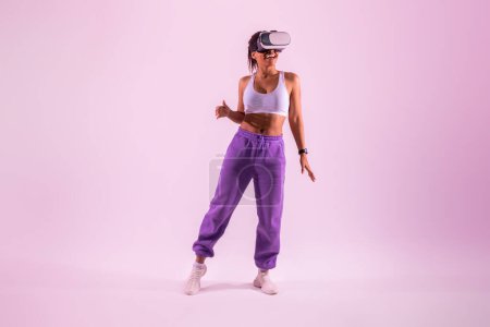 Téléchargez les photos : Heureuse femme noire dansant dans un casque de réalité virtuelle, profitant de son expérience de réalité virtuelle tout en explorant la technologie 3D sur fond de lumière rose néon, pleine longueur - en image libre de droit