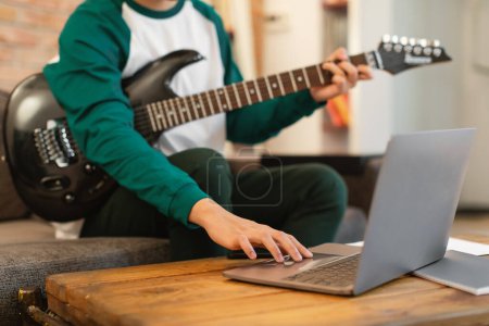 Téléchargez les photos : Enfant méconnaissable jouant de la guitare électrique à l'aide d'un ordinateur portable écrivant une chanson assis à la maison le week-end. Hobby et instruments de musique Concept. Cultivé, Focus sélectif - en image libre de droit