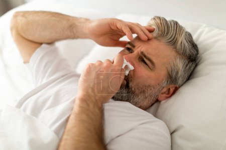 Closeup shot of sick middle aged grey-haired man wearing pajamas touching runny nose with napkin, sneezing in bed, touching his head, suffering from cold, flu, coronavirus