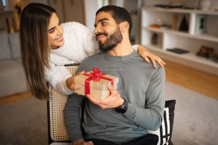 Photo for Glad young caucasian woman congratulates middle eastern male, gives box with present in room interior. Surprise for birthday, anniversary, holiday, best idea for gift, celebrate Valentine day at home - Royalty Free Image