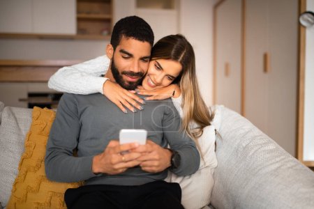 Photo for Smiling millennial european woman hugs arab husband plays game on smartphone, reads message in living room interior. Love, relationships, app and social network, communication remote with device - Royalty Free Image