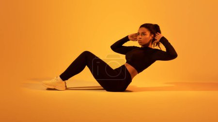 Téléchargez les photos : Jeune femme afro-américaine faisant des exercices abs sur fond de studio de lumière au néon orange, panorama, espace libre. Prise de vue pleine longueur de dame en forme travaillant sur les muscles de base - en image libre de droit