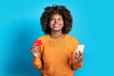 Photo for Emotional cheerful attractive young black woman with bushy hair and teeth braces holding red bank card and cell phone in her hands, purchasing online, blue studio background - Royalty Free Image