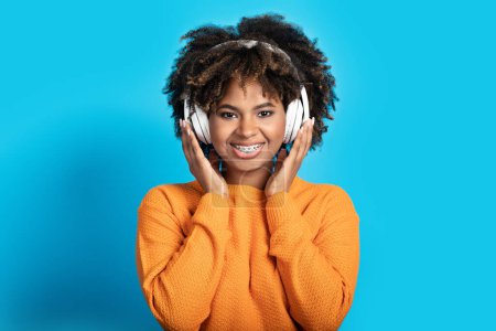 Photo for Closeup of positive cheerful pretty millennial black lady with bushy hair and teeth braces using wireless headphones, look at camera and smiling on blue studio background, enjoying sound - Royalty Free Image