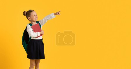 Photo for Look There. Cute Little Schoolgirl Pointing Finger Aside At Copy Space While Standing Isolated Over Yellow Background, Smiling Female Child Demonstrating Free Place, Advertising Something, Panorama - Royalty Free Image
