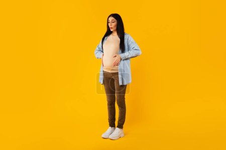 Photo for Full body length shot of pregnant woman suffering from abdomen pain, touching belly and breathing, standing on yellow studio background, copy space - Royalty Free Image