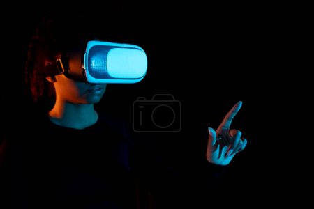 Photo for Young african american woman in VR goggles choosing options on invisible screen while having virtual reality experience in dark room with neon lights, copy space - Royalty Free Image