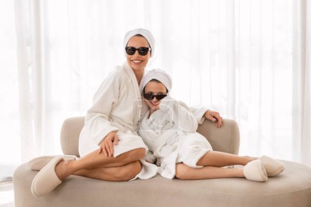Téléchargez les photos : Portrait de maman heureuse et sa petite fille portant des peignoirs et des lunettes posant à la caméra tout en se relaxant sur le canapé à la maison, famille joyeuse s'amuser et profiter de la journée de beauté, Copier l'espace - en image libre de droit