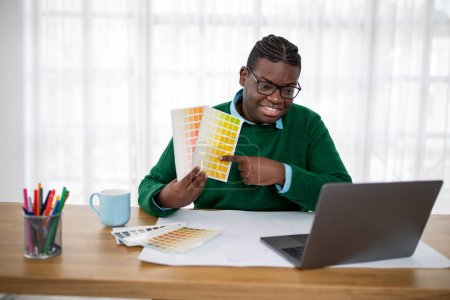 Foto de Alegre hombre afroamericano diseñador de elección de color en la paleta que lo muestra a la computadora portátil videollamadas Comunicarse y trabajar en línea en la oficina moderna. Concepto de diseño profesional - Imagen libre de derechos